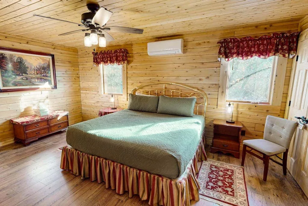 Dormitorio con una cama, paredes revestidas de madera y ventanas con cortinas estampadas.