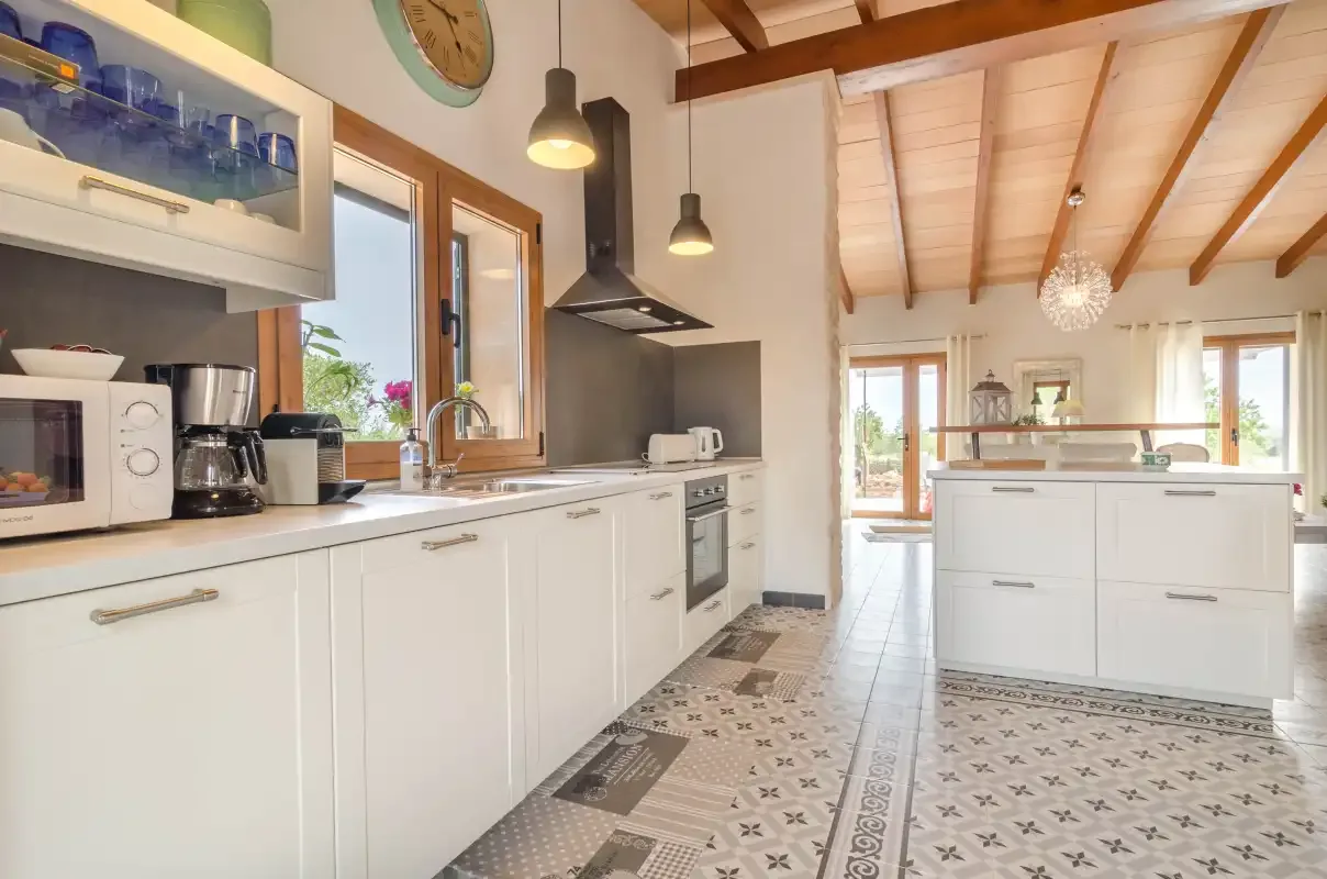 Una cocina luminosa con armarios blancos, una isla central y vigas vistas en el techo.