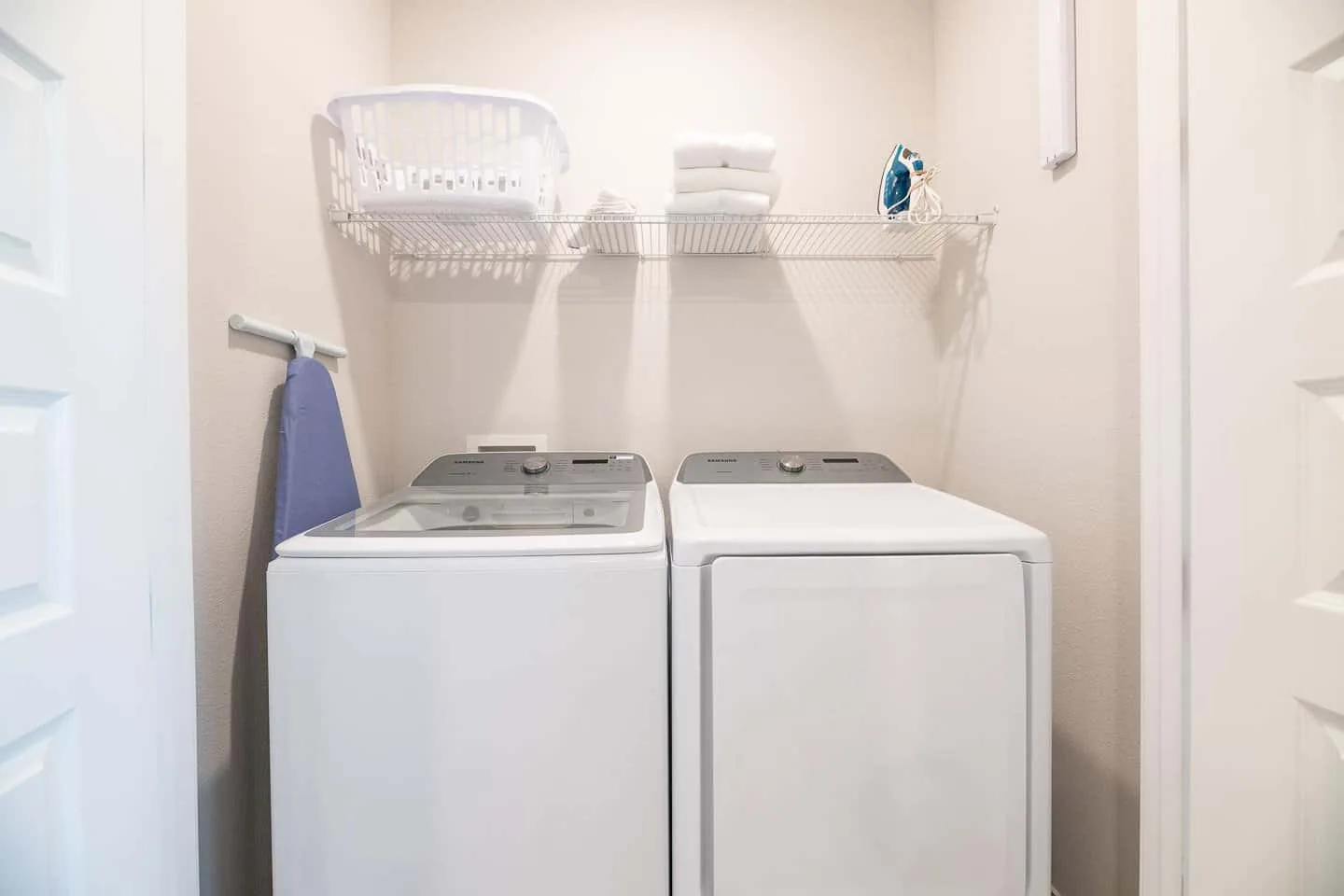 A laundry room with a top-loading washer and dryer beneath a wire shelf.
