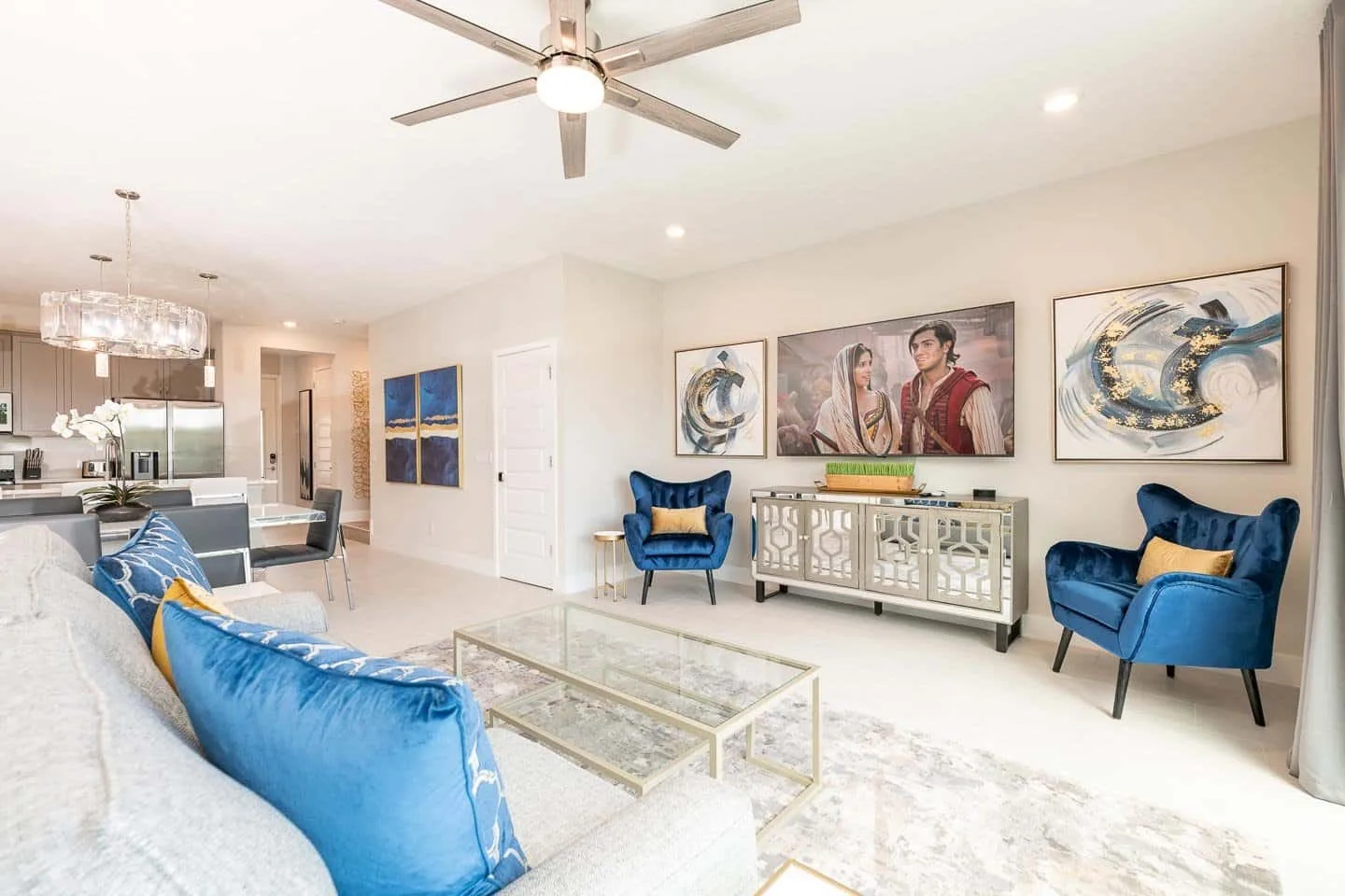 A bright living room has blue seating, a glass coffee table, and framed artwork.