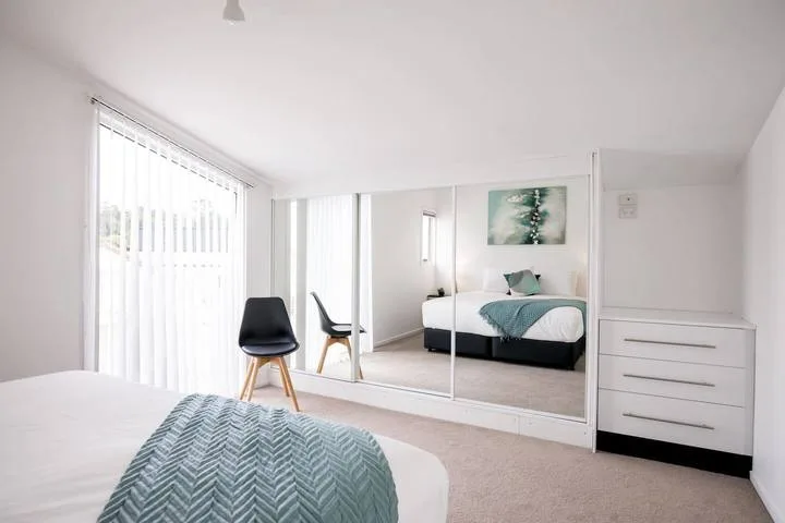A bright bedroom with a bed, mirrored wardrobe, and chair beside the window.