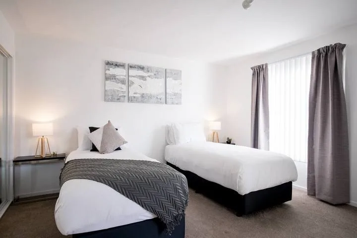 A bright bedroom with two beds, bedside lamps, and a curtained window.