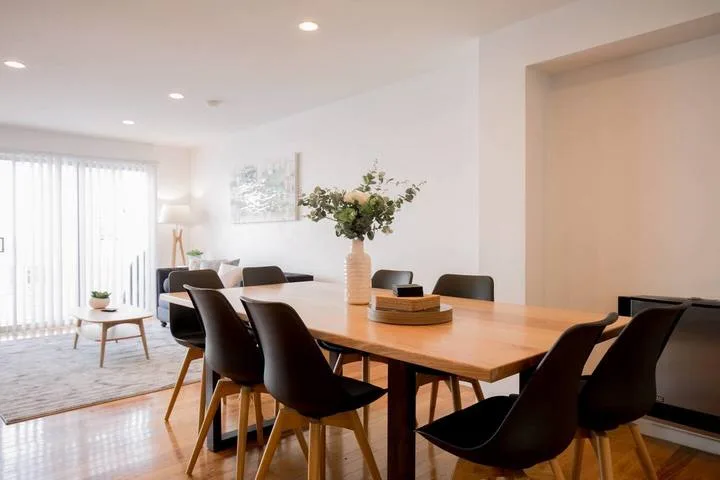A bright dining area features a wooden table, dark seating, and flowers.