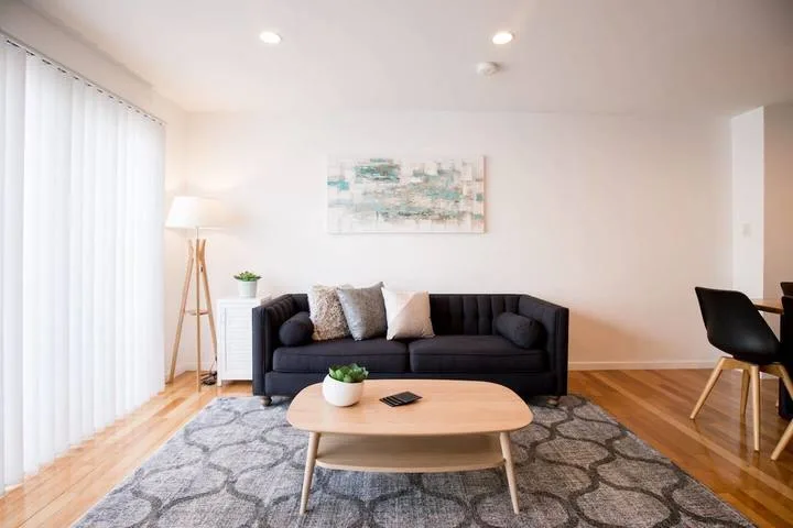 A bright living room with dark seating, a coffee table, and a patterned rug.