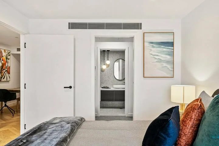 A bright bedroom with a bed, colorful pillows, and an open doorway to a bathroom.