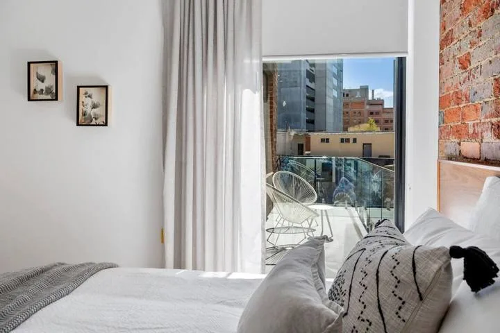 A bright bedroom with white bedding and a large window overlooking nearby buildings.