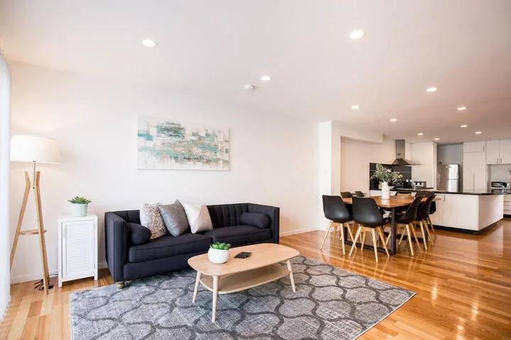 A bright open-plan living and dining area with dark seating and a kitchen beyond.