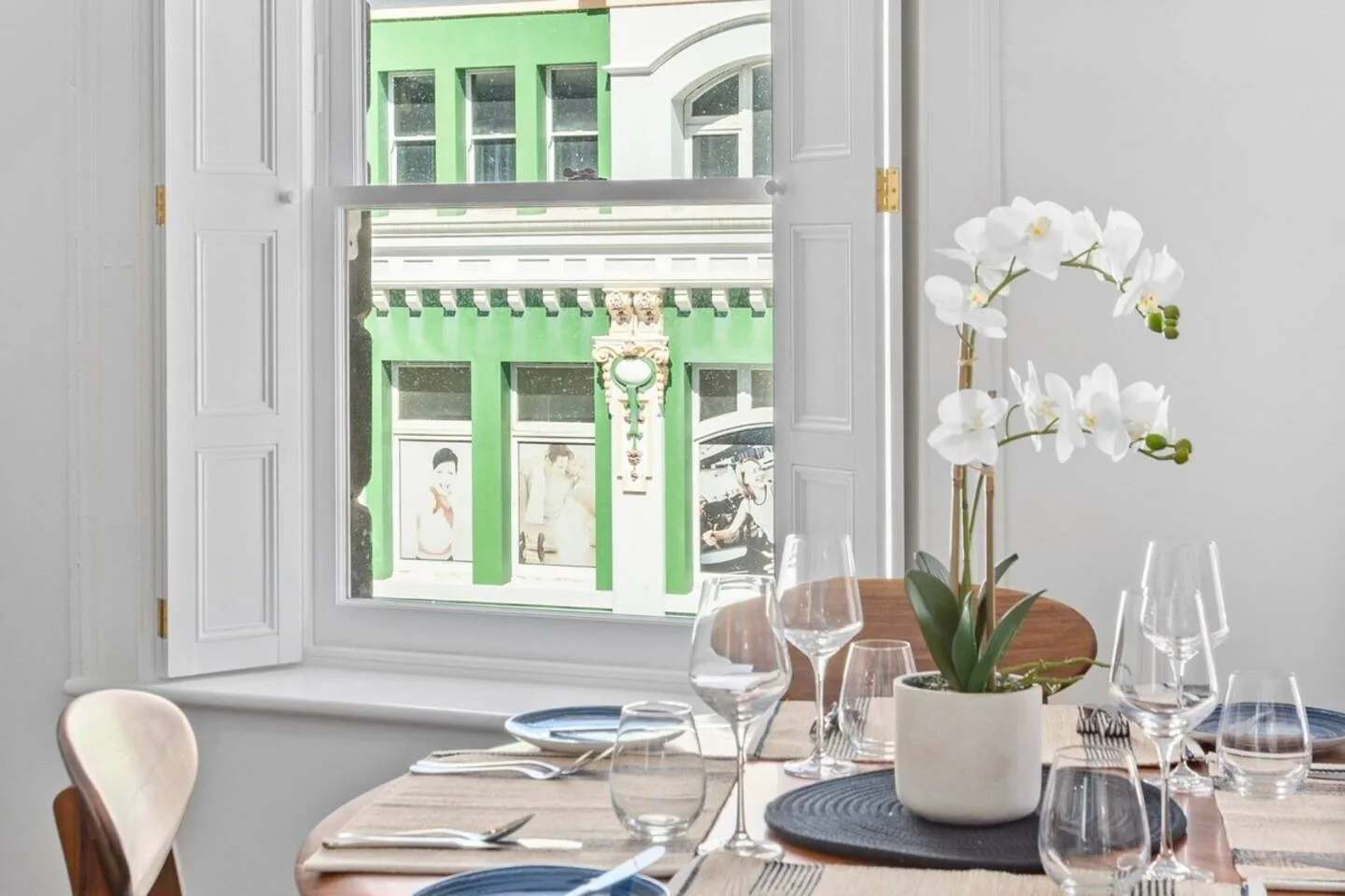 A dining table set with glassware sits beside open windows overlooking a green building.