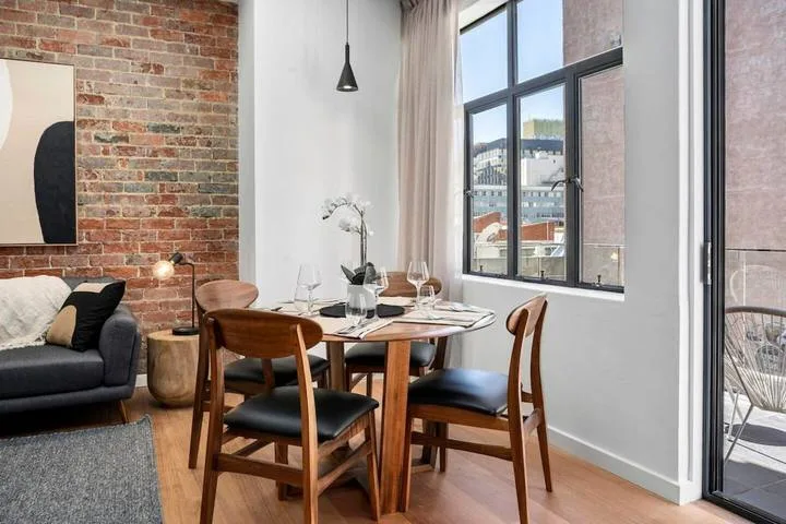 A dining area with a wooden table and chairs beside large windows.