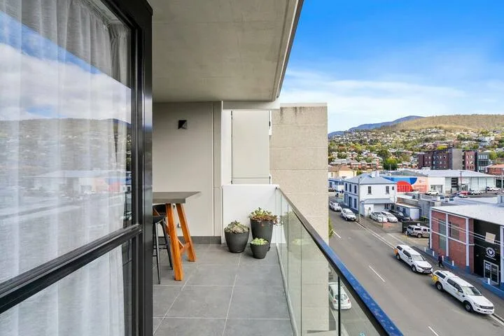A balcony overlooks a city street, with potted plants beside the glass railing.