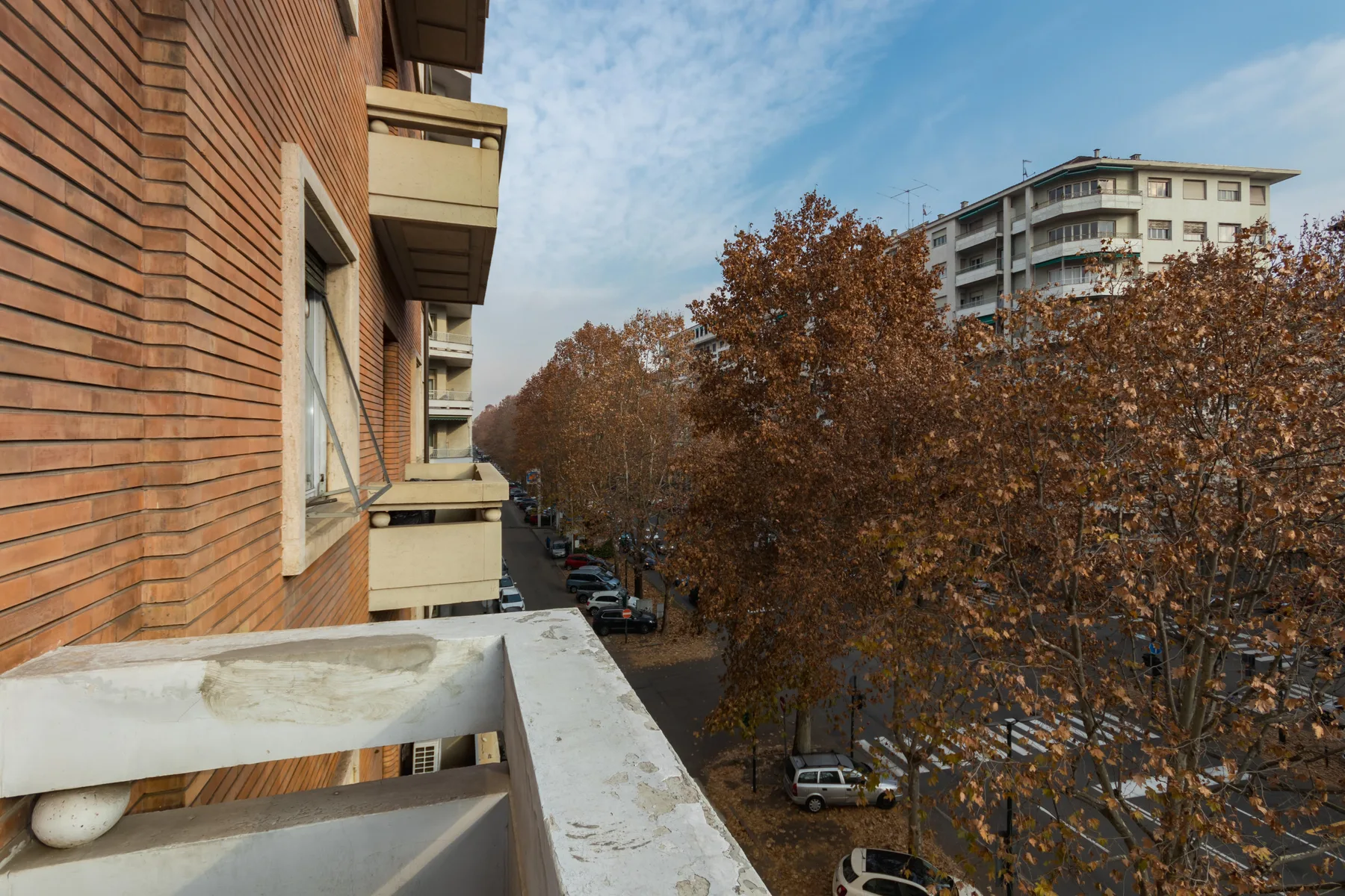 View from a balcony overlooking a tree-lined street and nearby apartment buildings.