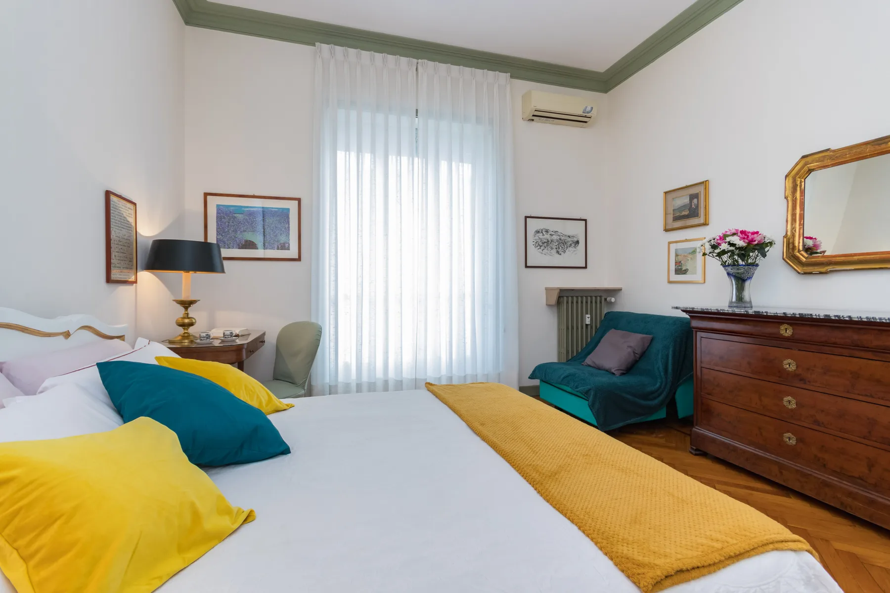 A bedroom with a large bed, a teal armchair, and a tall curtained window.