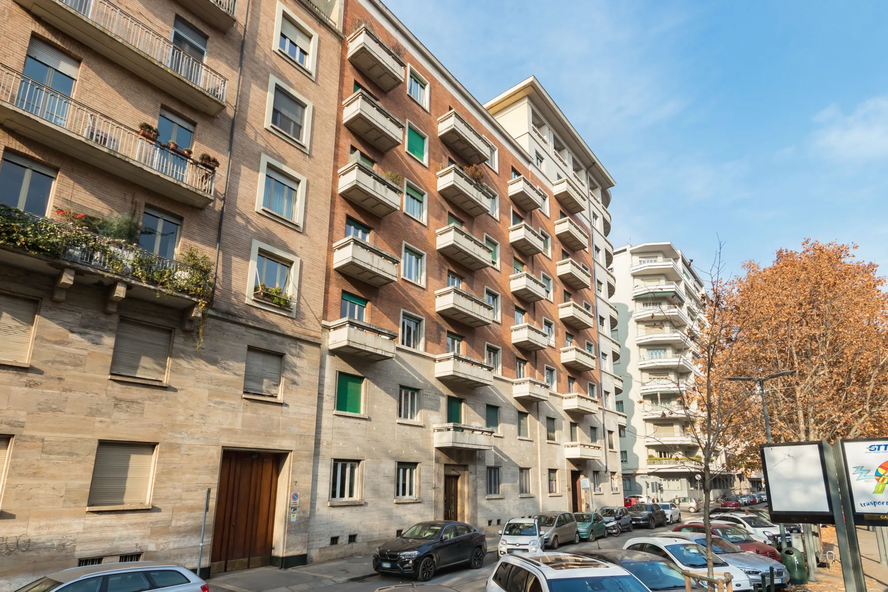 Escena de calle urbana con edificios de viviendas, coches aparcados y árboles otoñales.