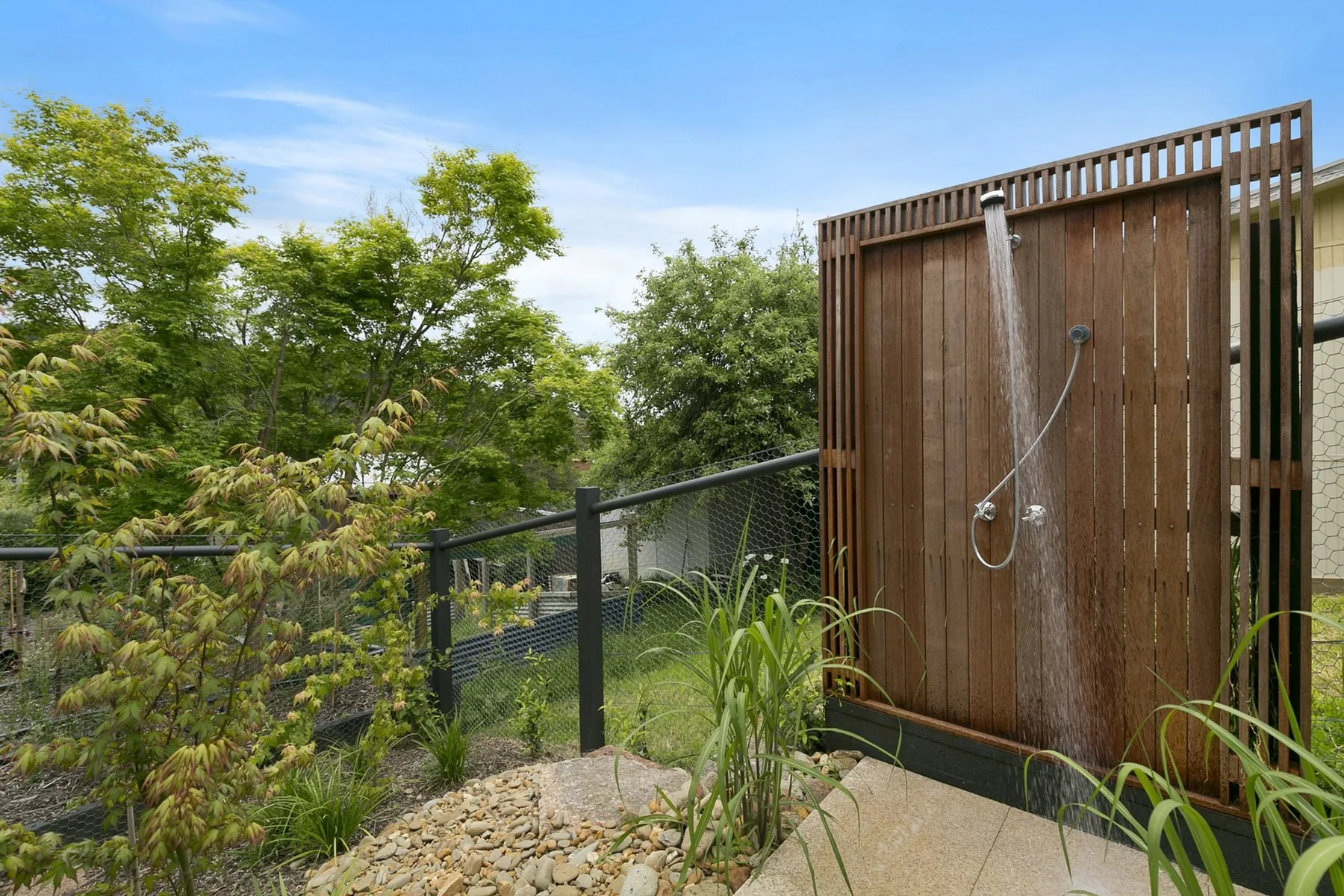 An outdoor shower beside a wooden screen and garden greenery.