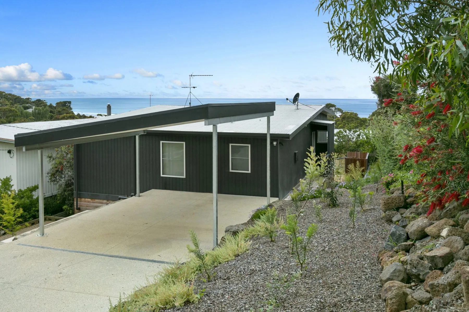 A modern dark house with a covered driveway sits beside a gravel garden.