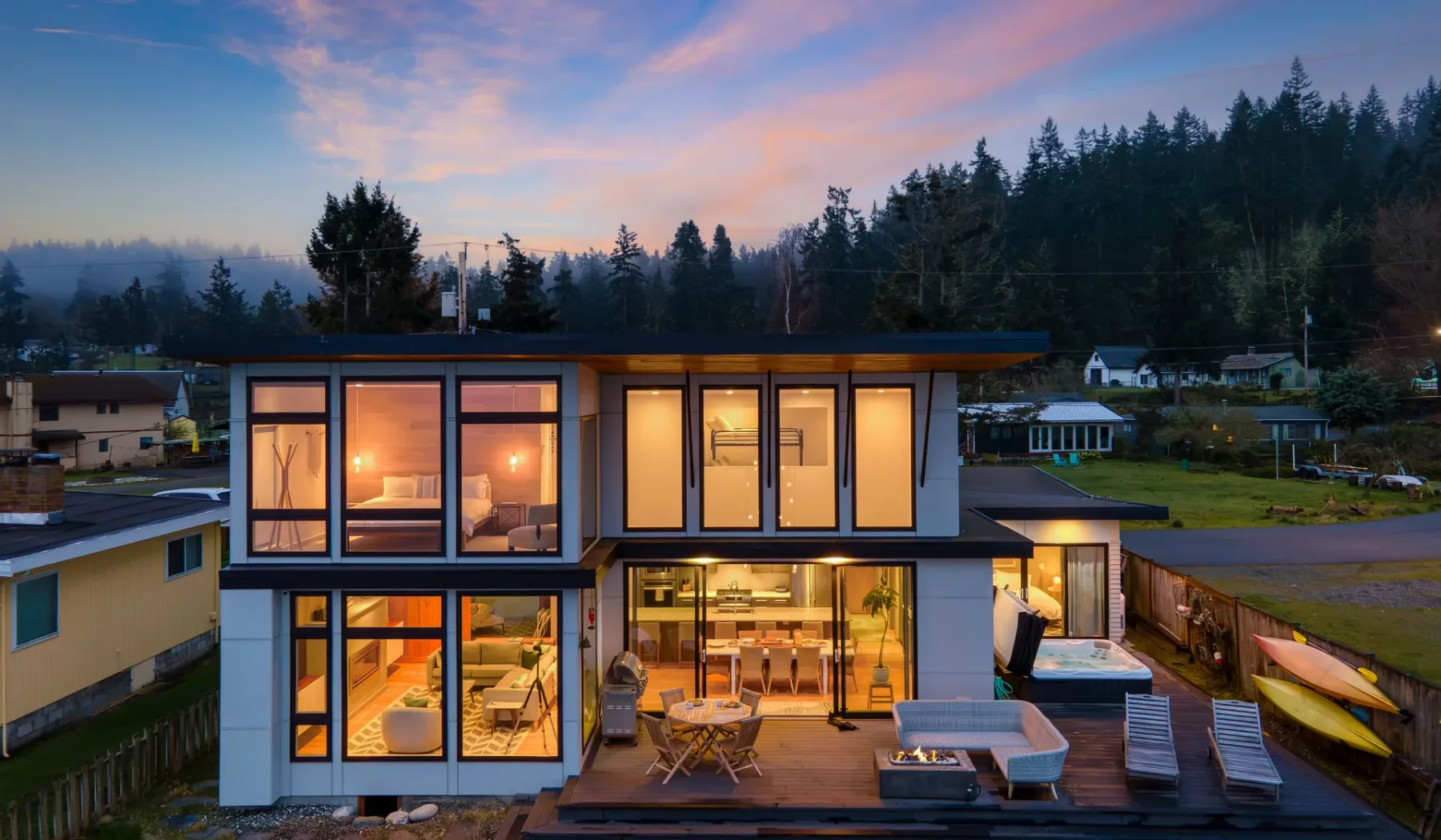 A modern two-story house glows at dusk, with a forested hillside behind it.