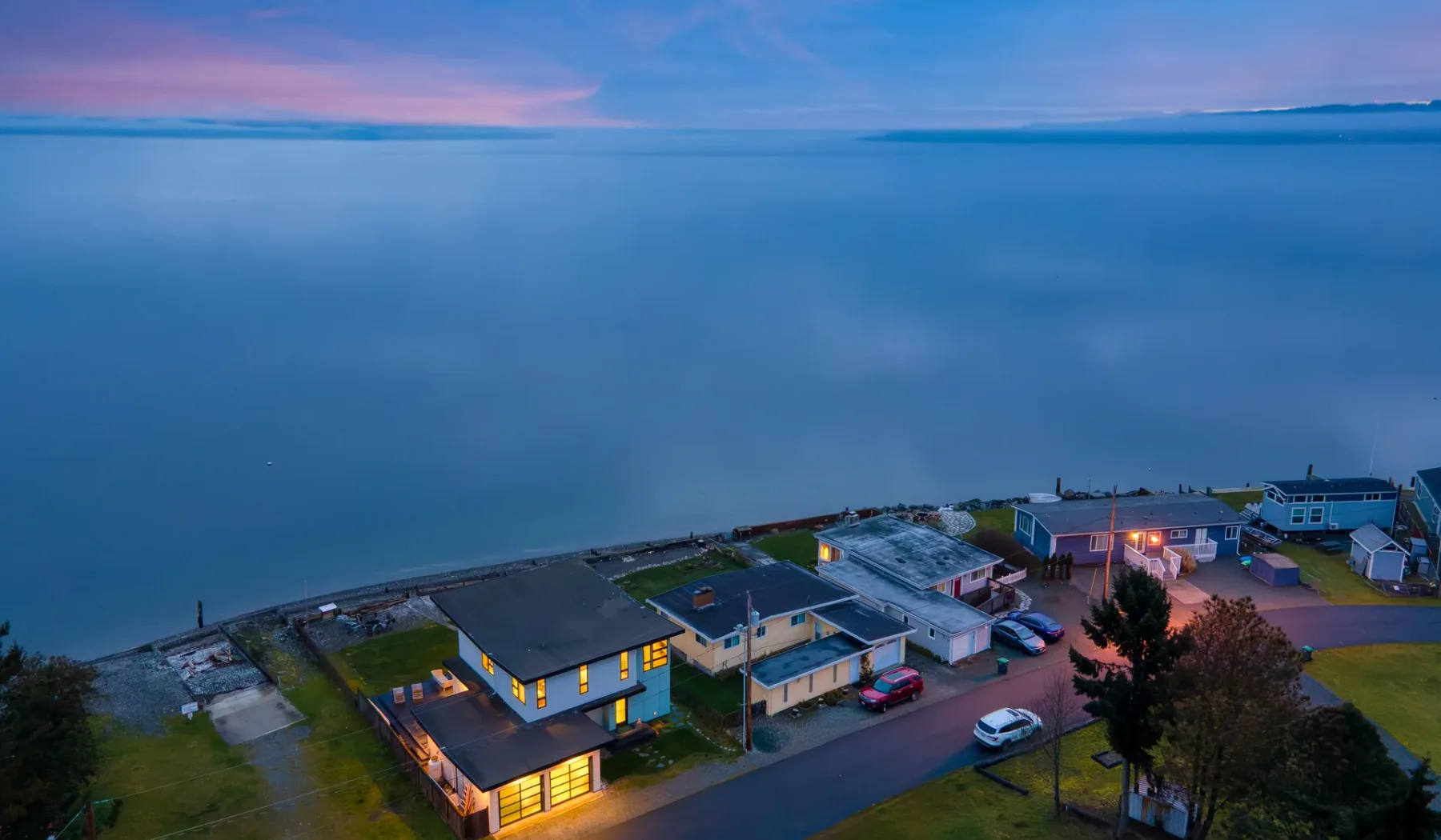 A waterfront building and nearby homes sit beside a calm bay at dusk.