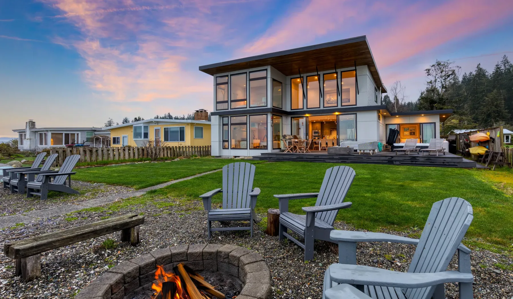 A modern house overlooks a grassy yard with outdoor seating and a fire pit.