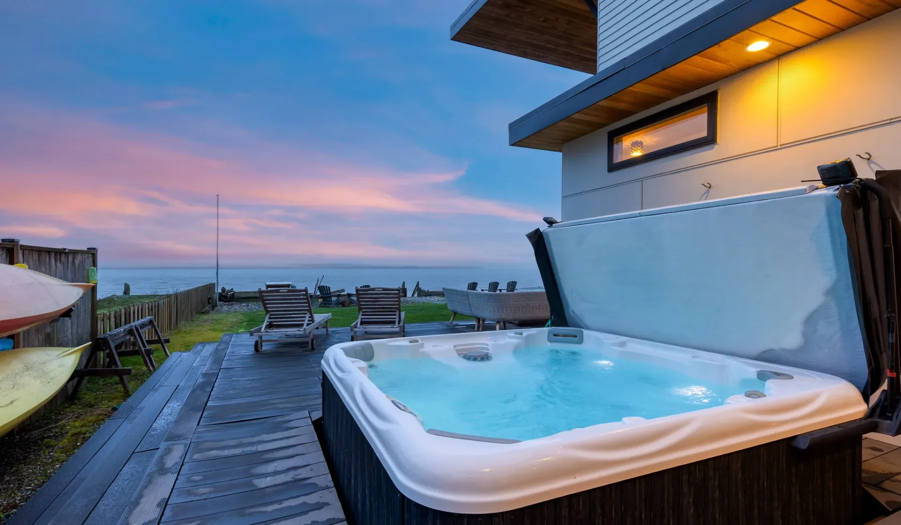 An outdoor hot tub beside a wooden deck overlooks the water at sunset.