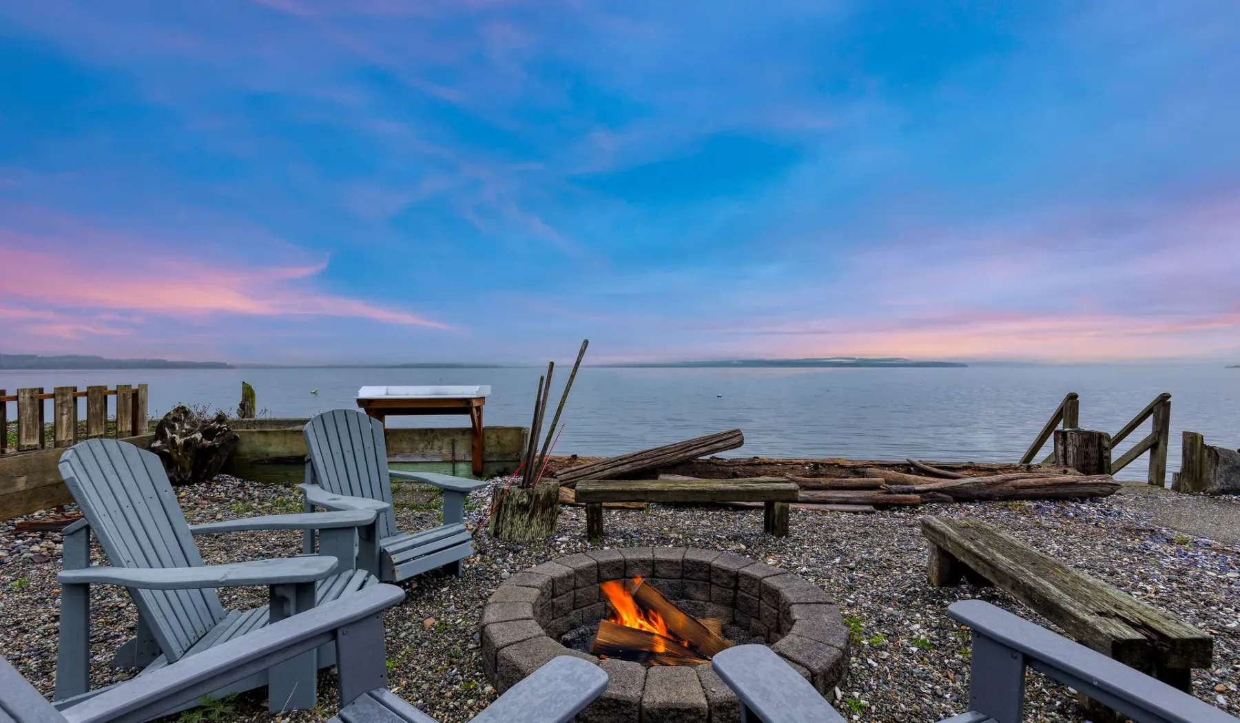 A lakeside fire pit with outdoor seating overlooks calm water at sunset.