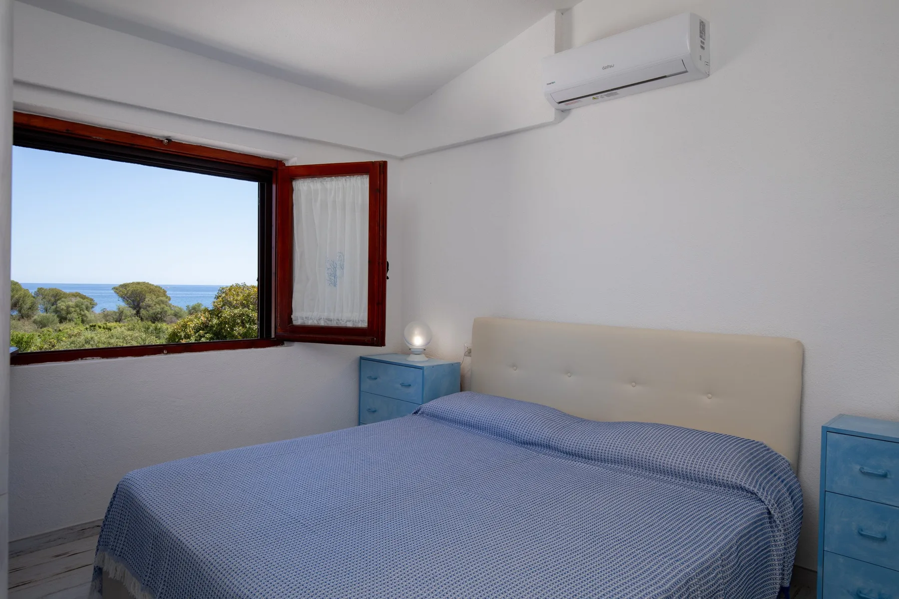 A bedroom with a bed beside an open window overlooking greenery and the sea.