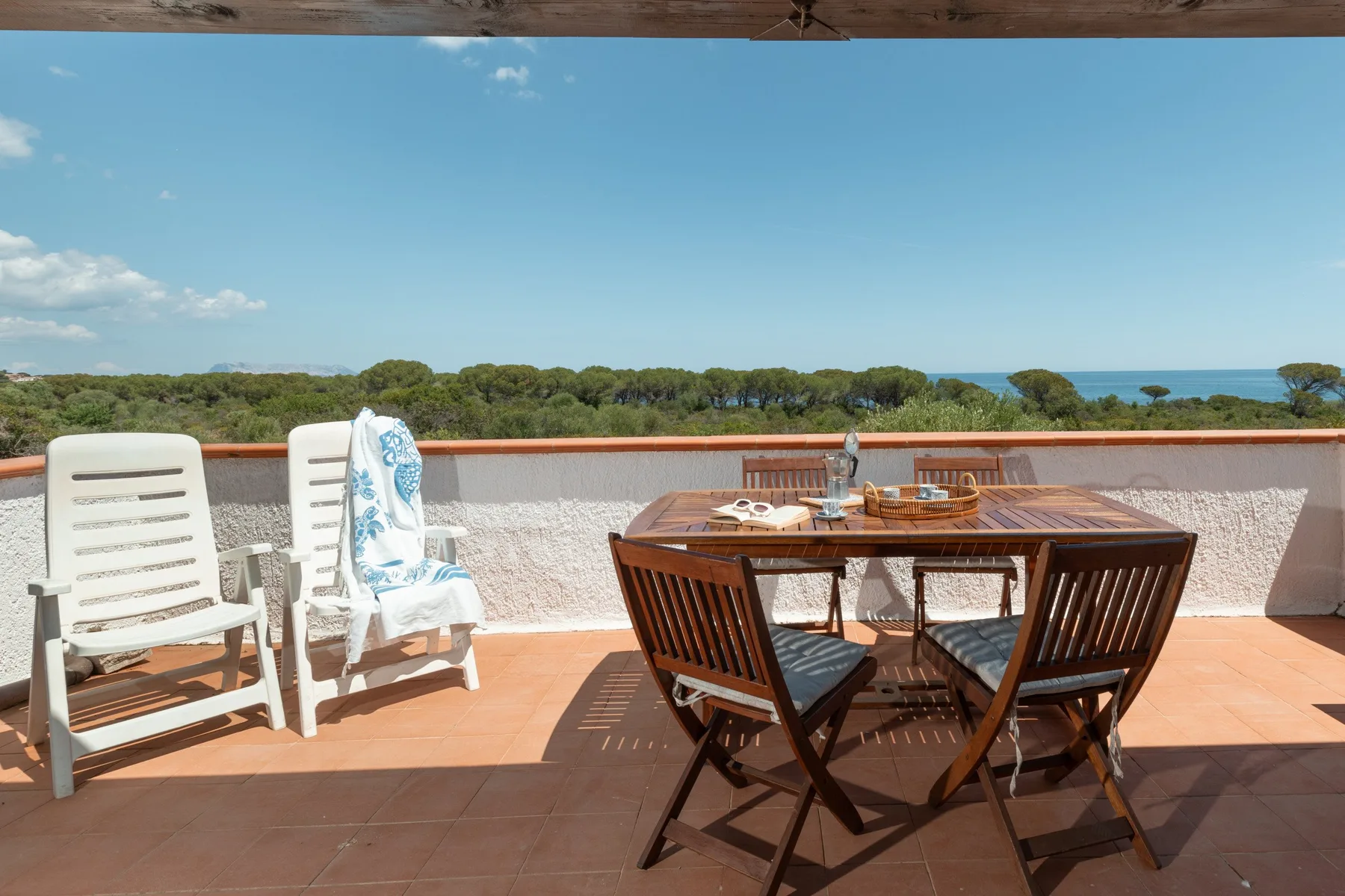 A sunny terrace with outdoor seating and a dining table overlooking trees and the sea.