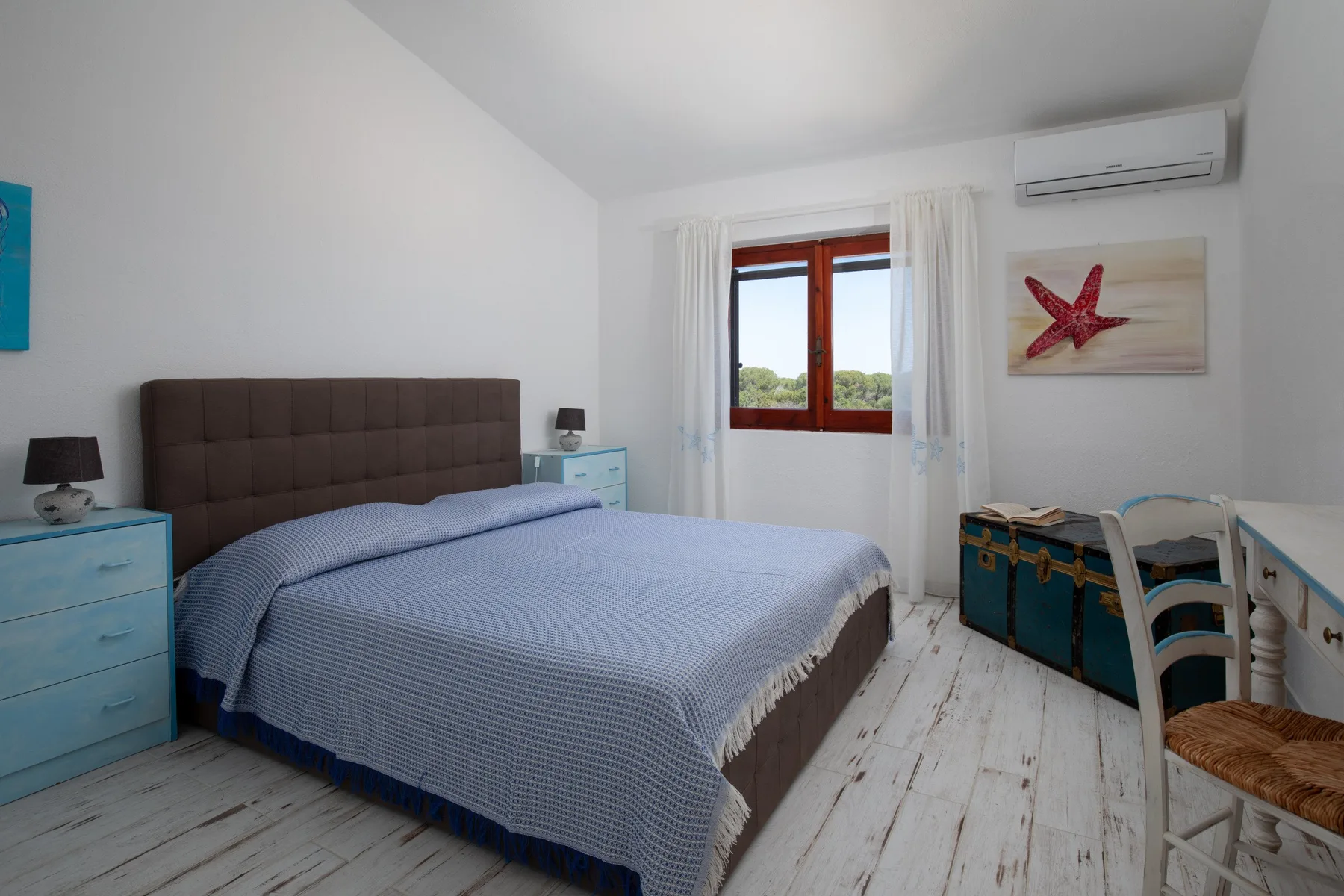 A bedroom with a bed, blue bedside furniture, and a window with white curtains.