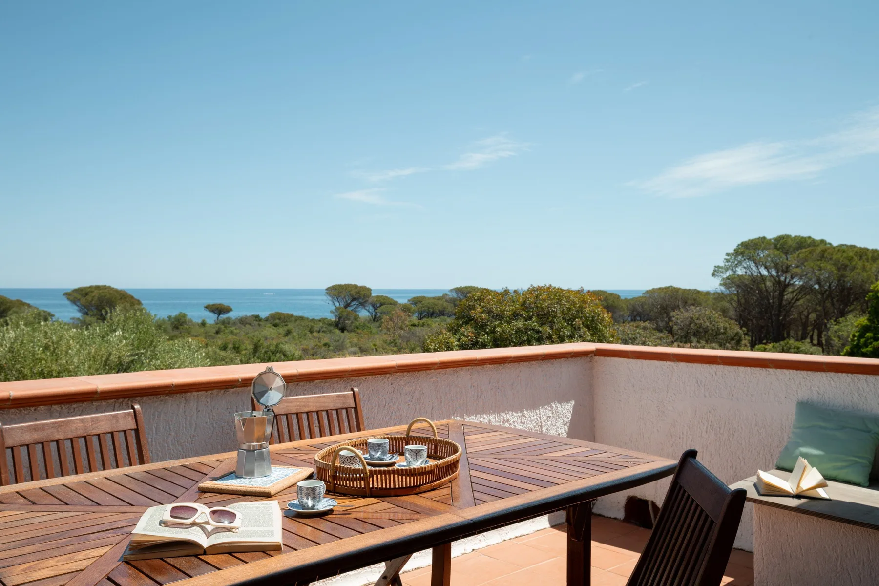 A rooftop terrace with a wooden dining table overlooks greenery and the sea.