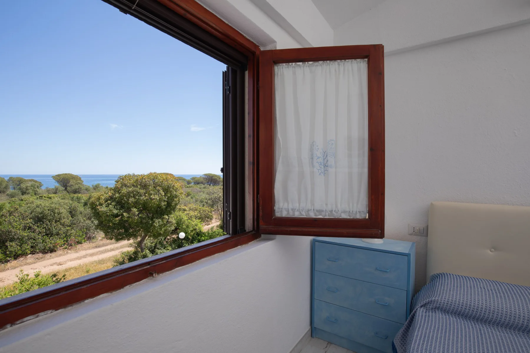 A bedroom with an open window overlooking greenery and the sea.