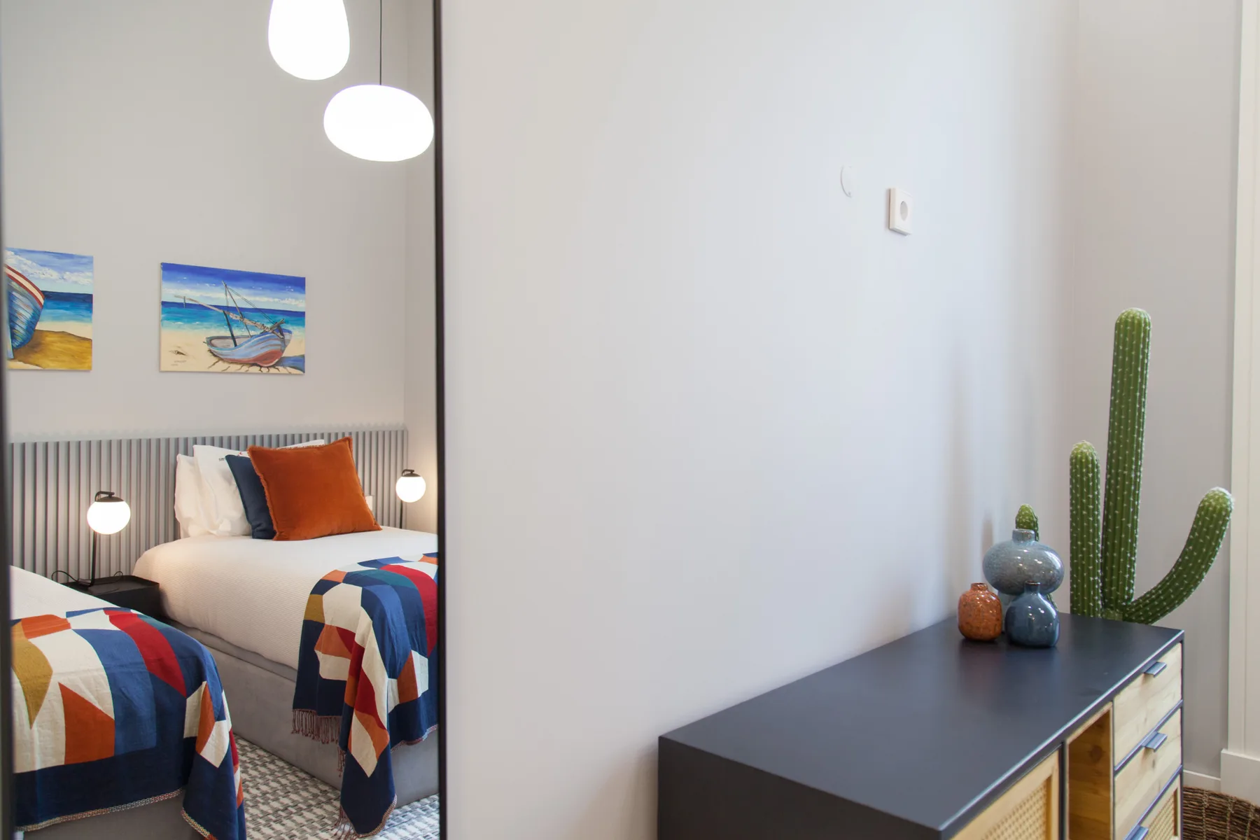 A bright bedroom with colorful bedding, wall art, and a cactus on a console.