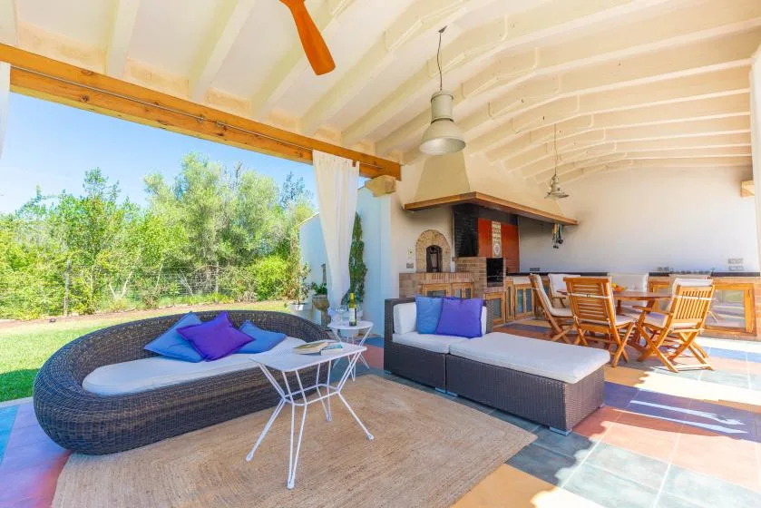 Un patio cubierto tiene asientos al aire libre junto a una mesa de comedor y vegetación alrededor.