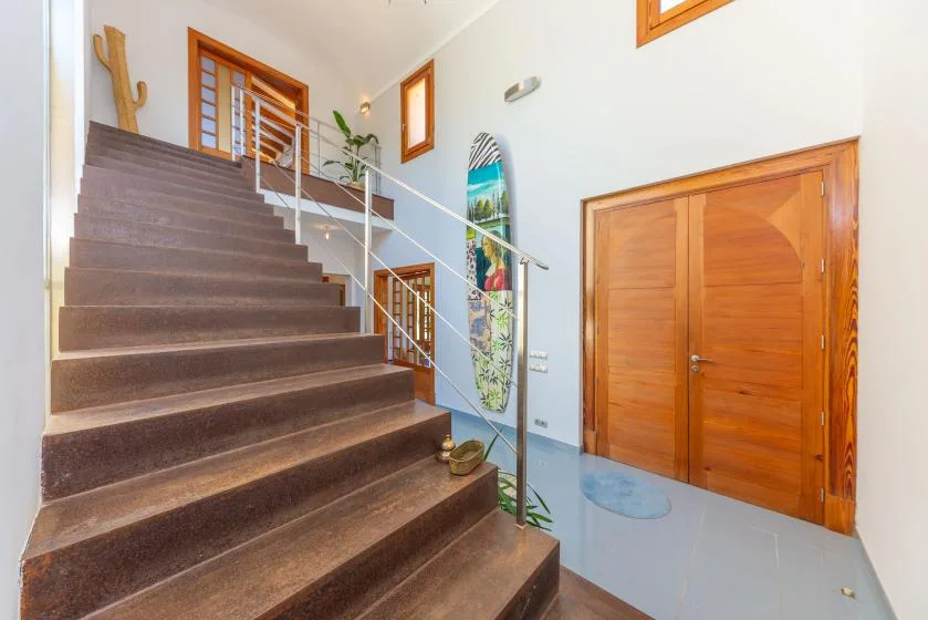 Un recibidor luminoso con una escalera, barandilla de vidrio y puertas de madera.