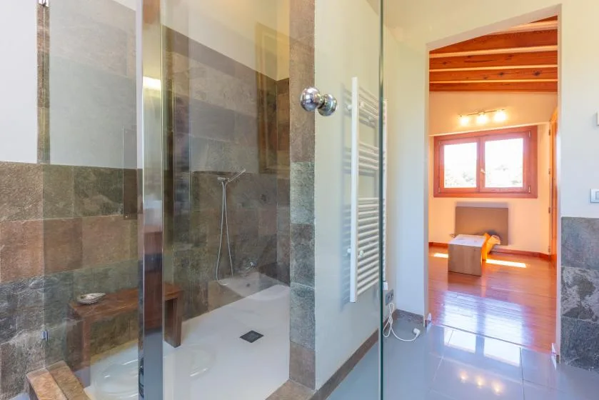 Un baño con ducha de vidrio y una puerta abierta a una habitación luminosa contigua.