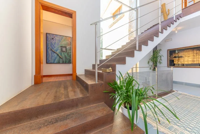 Una escalera interior luminosa con barandilla de vidrio y una planta en maceta junto a los escalones.