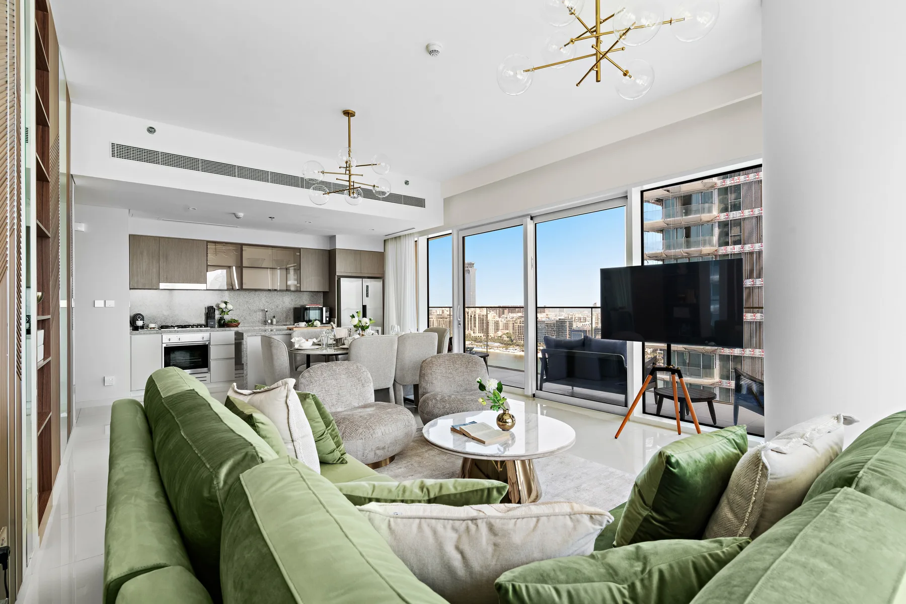 A bright open-plan living room with green seating, a dining area, and large windows.