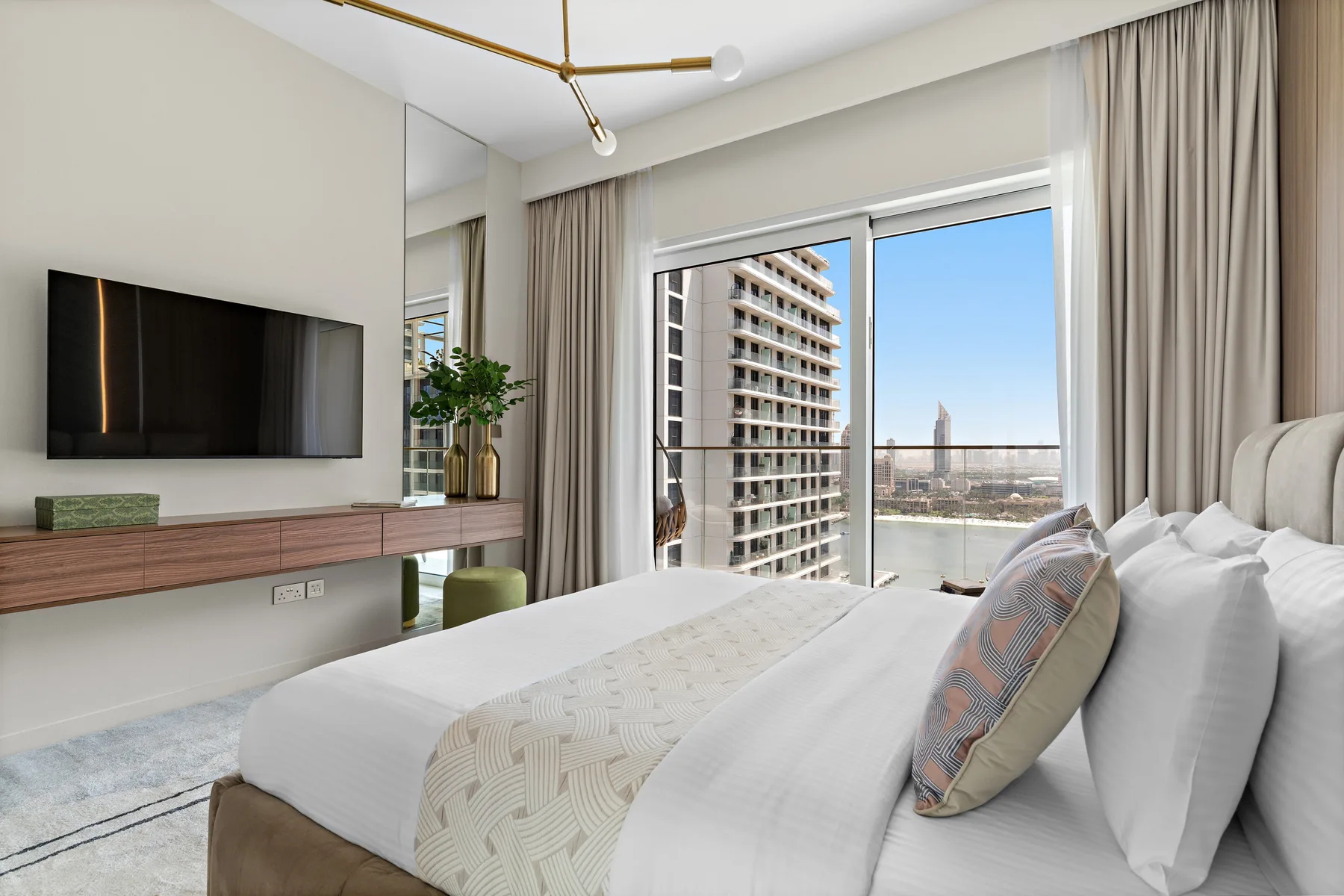 Un dormitorio luminoso con una cama tendida junto a grandes ventanas con vistas a edificios cercanos.