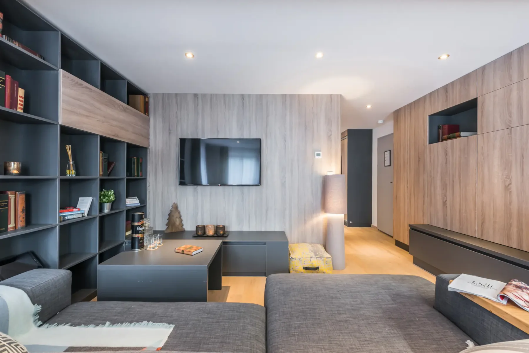 Un salón moderno con asientos oscuros, estanterías empotradas y un televisor en la pared.