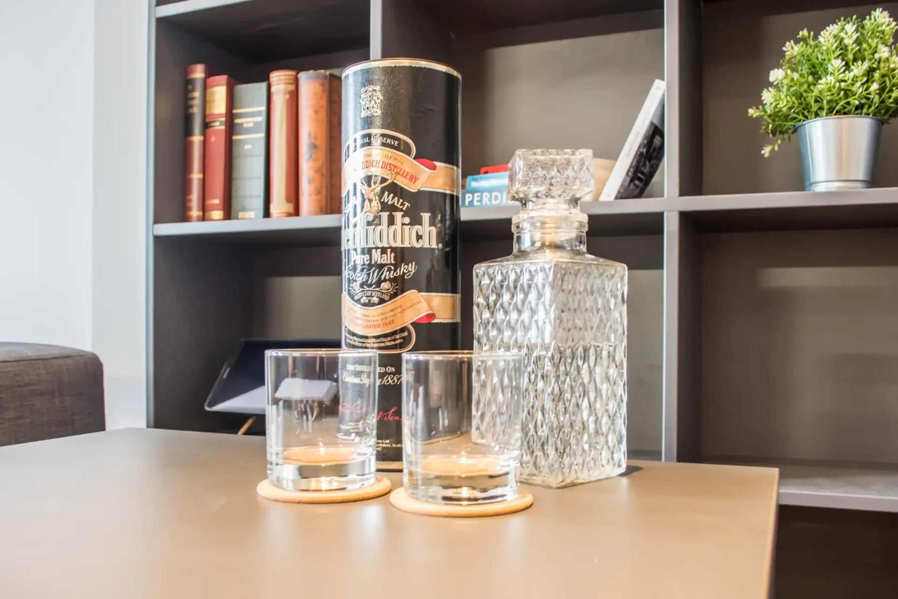 Una mesa muestra una botella de whisky, dos vasos y una licorera de vidrio.
