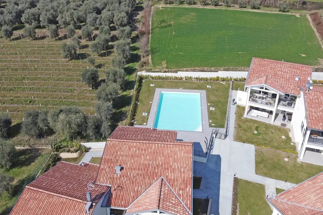 An aerial view shows a swimming pool beside tiled-roof buildings and green fields.
