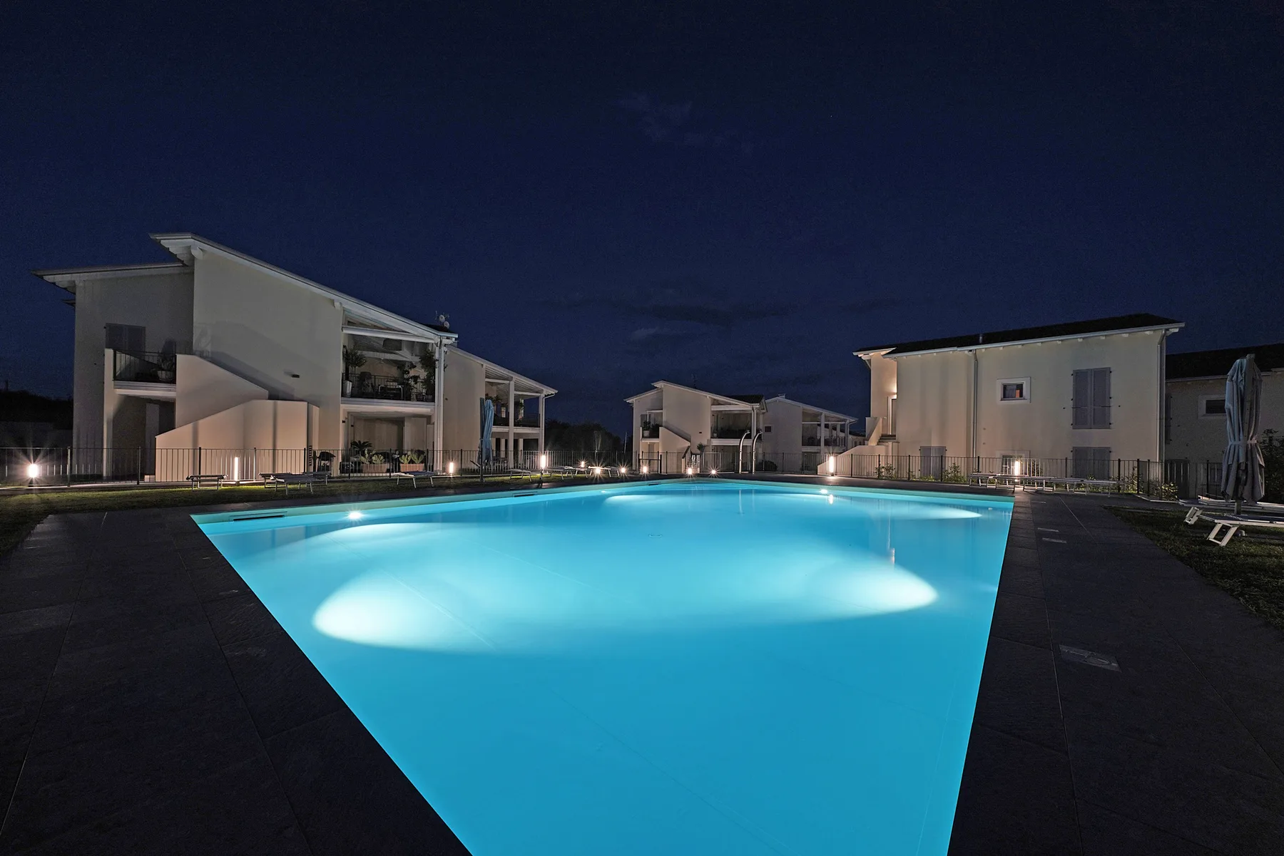 A brightly lit outdoor swimming pool sits among modern buildings at night.
