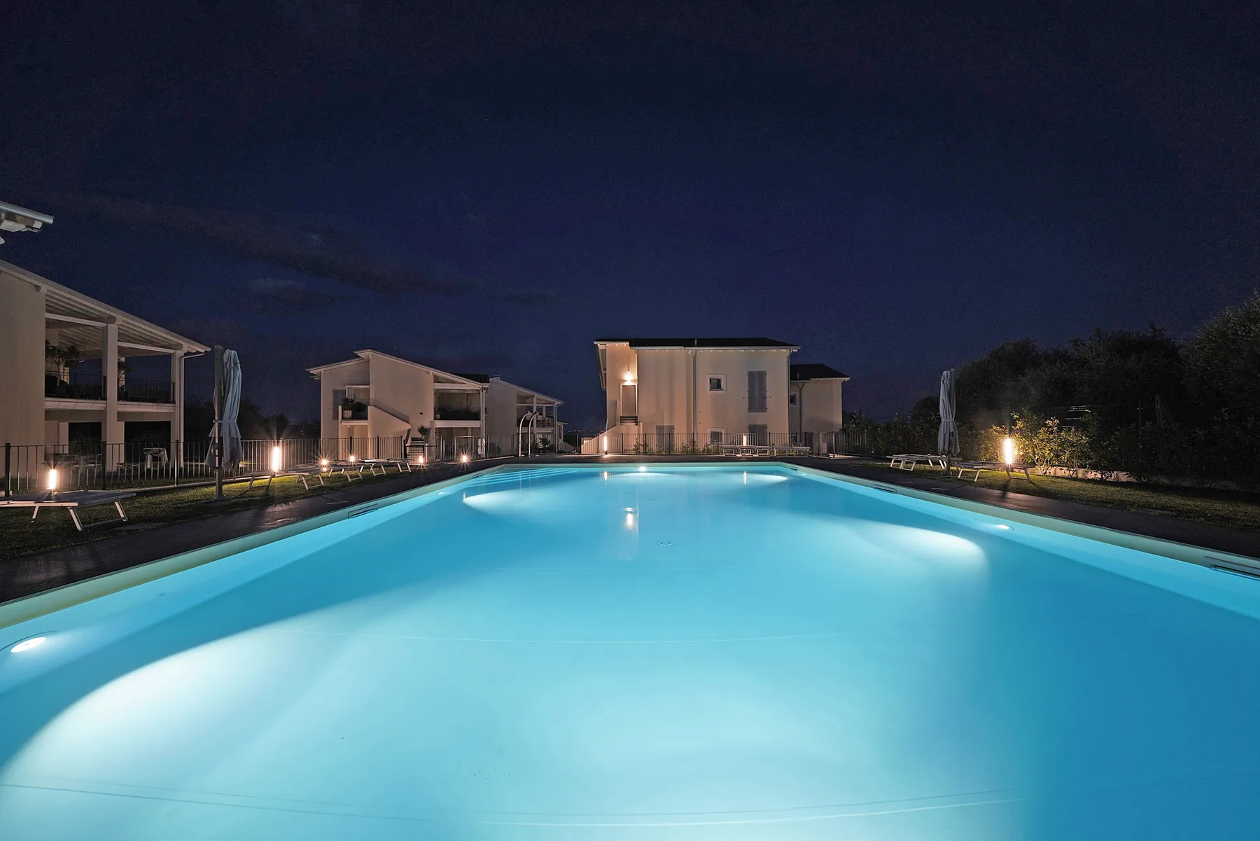 An illuminated swimming pool at night, with buildings along the far side.
