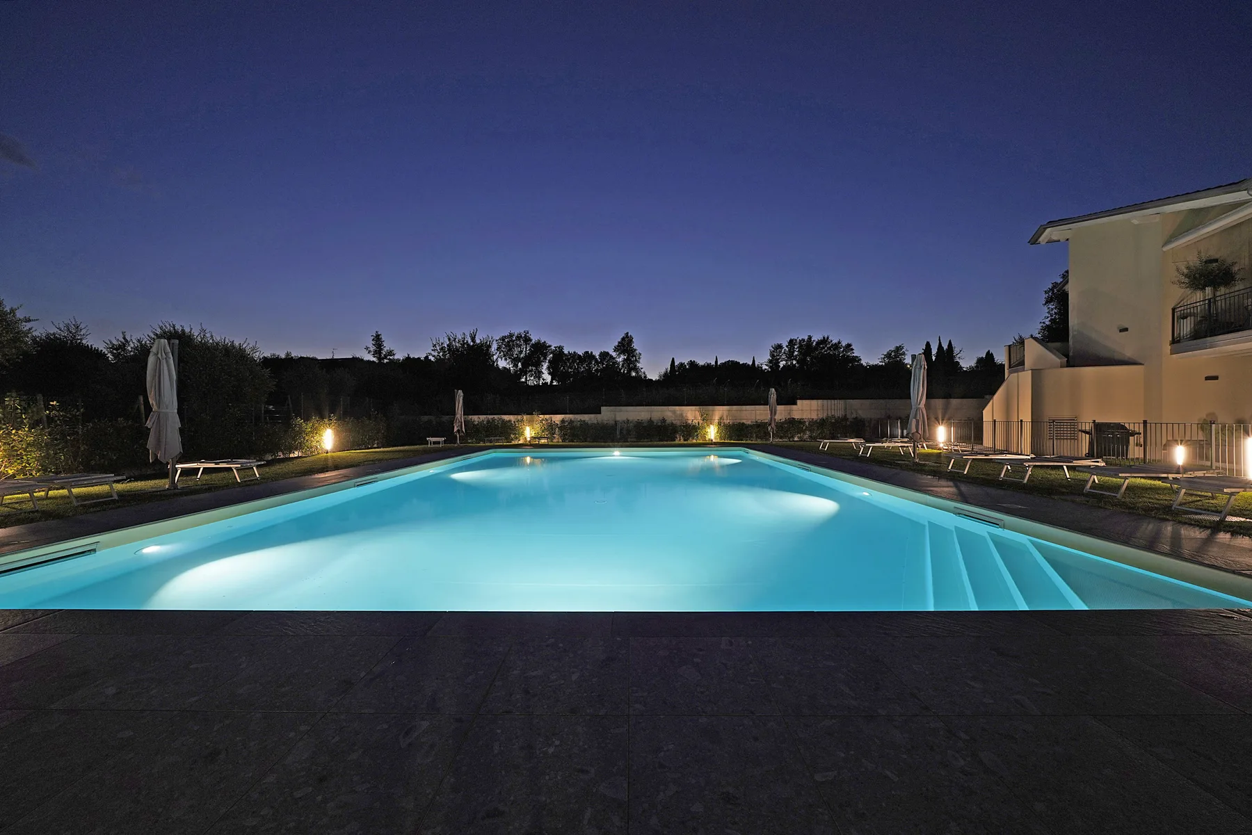 An illuminated outdoor swimming pool at dusk, with lounge chairs along its sides.
