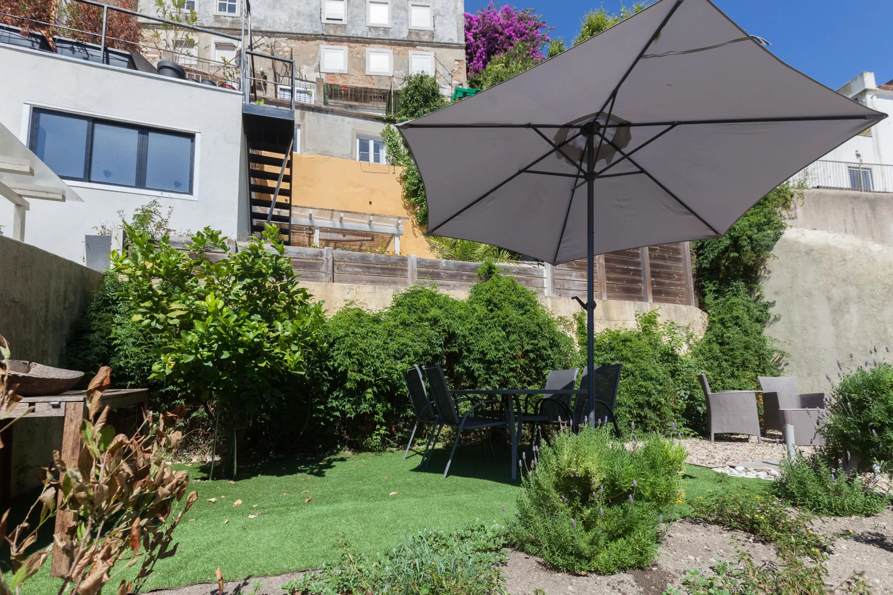 Una zona de estar en el jardín tiene una sombrilla grande y vegetación alrededor.