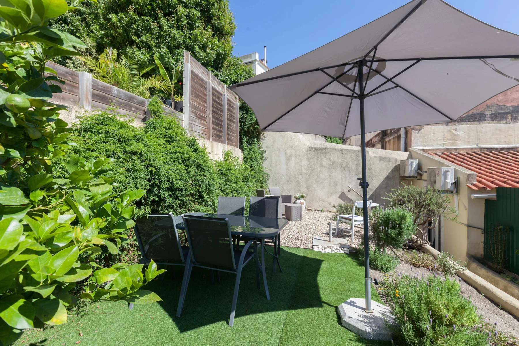 Un patio con asientos al aire libre está bajo una sombrilla grande, rodeado de vegetación.