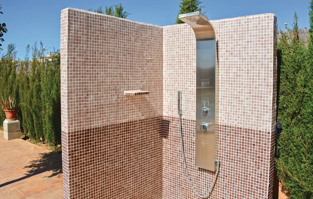 Una ducha exterior de azulejos está junto a un seto en un patio soleado.