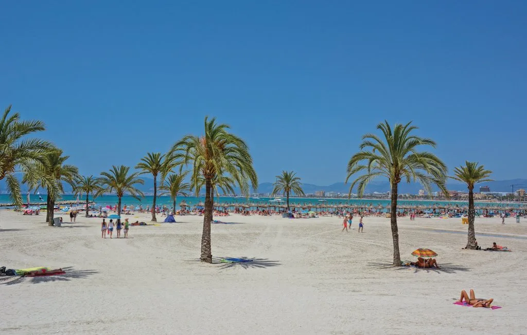 Una playa de arena con palmeras, bañistas y el mar al fondo.