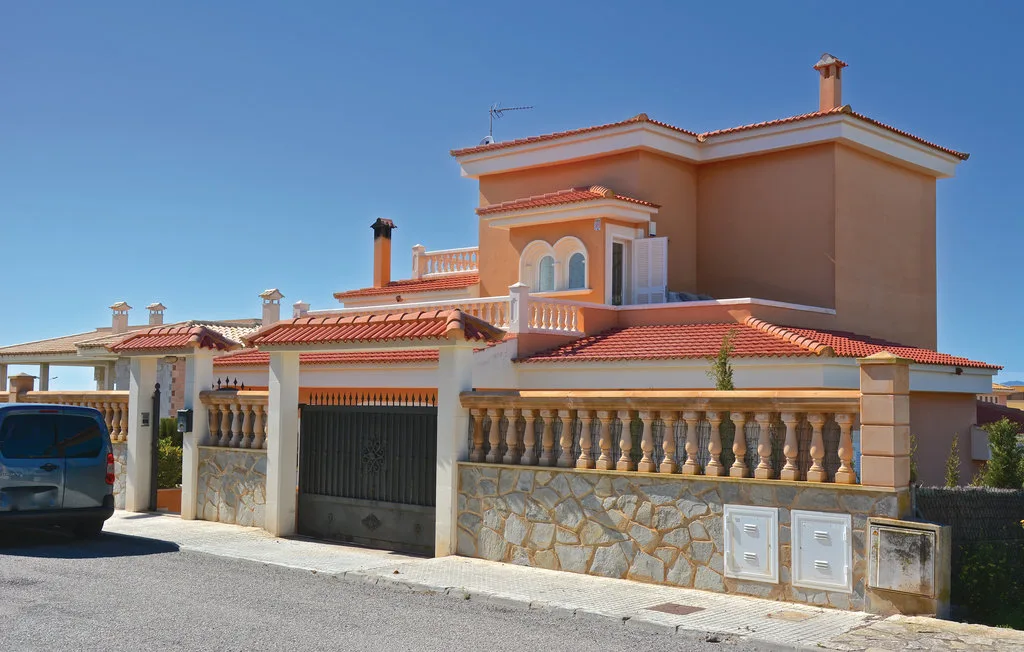 Una casa grande de estuco se alza detrás de un muro de piedra y una entrada con portón.
