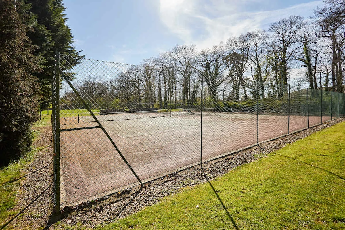 An outdoor tennis court is enclosed by a chain-link fence, with trees beyond.