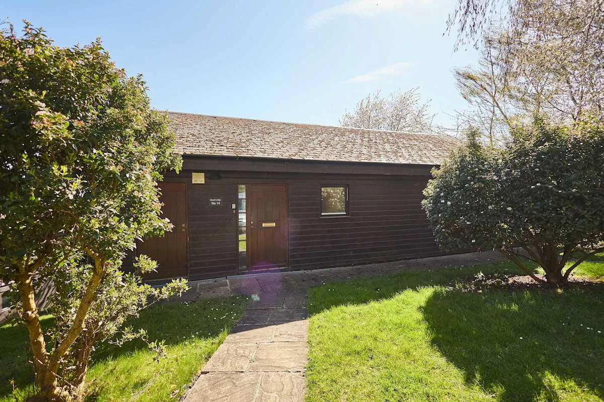 A dark wooden lodge with a central door stands beside a sunlit garden path.