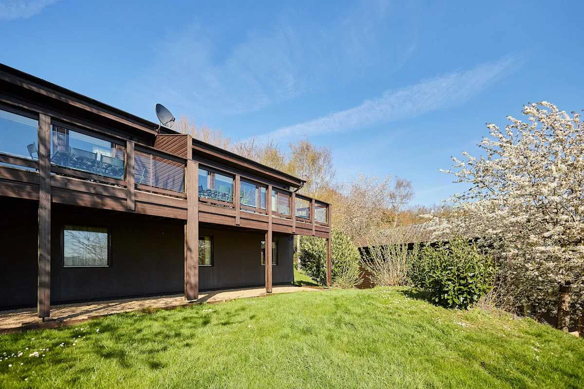 A dark wood lodge overlooks a grassy lawn, with trees and shrubs nearby.